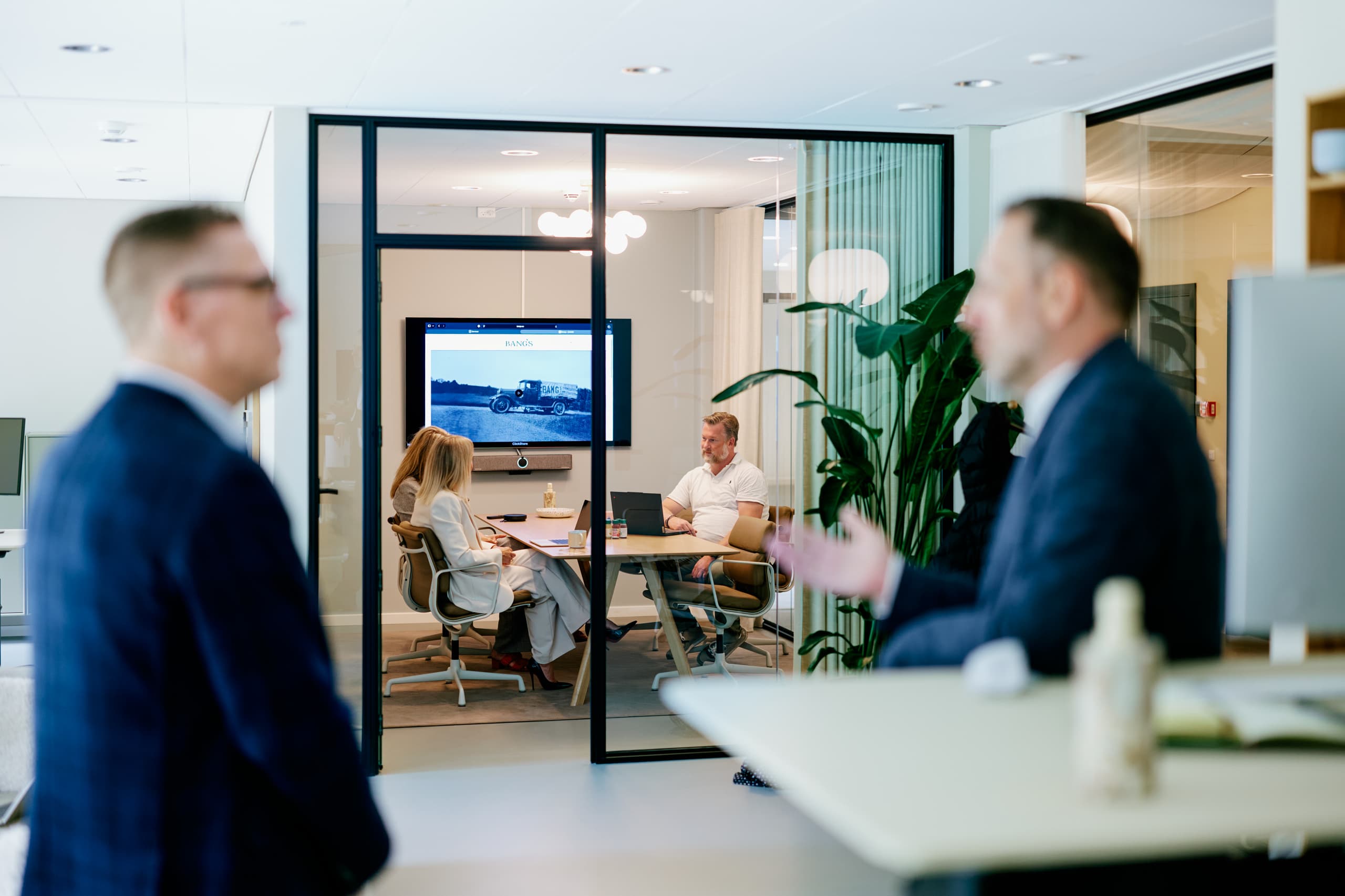 Zwei Männer im Gespräch in einem Büro, während im Glas-Besprechungsraum dahinter ein Meeting stattfindet